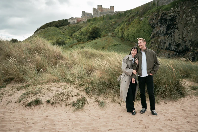 An engaged couple standing together among the dunes beneath Bamburgh Castle